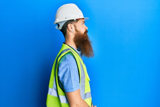 Redhead man with long beard wearing safety helmet and reflective jacket looking to side, relax profile pose with natural face with confident smile.