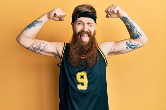Redhead man with long beard wearing basketball uniform showing muscles winking looking at the camera with sexy expression, cheerful and happy face.