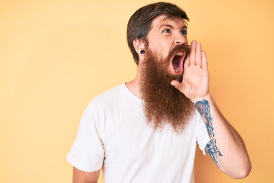 Handsome young red head man with long beard wearing casual white tshirt shouting and screaming loud to side with hand on mouth. communication concept.