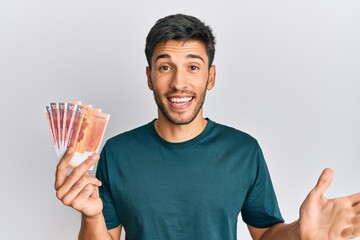 Young handsome man holding 100 norwegian krone banknotes celebrating achievement with happy smile...