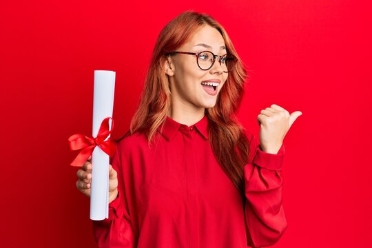 Young redhead woman holding graduate degree diploma pointing thumb up to the side smiling happy with open mouth