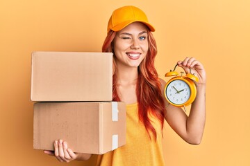 Young redhead woman holding delivery package and alarm clock winking looking at the camera with...