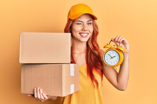 Young redhead woman holding delivery package and alarm clock winking looking at the camera with sexy expression, cheerful and happy face.