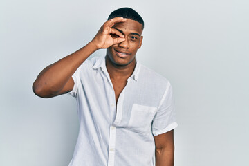 Young black man wearing casual white shirt doing ok gesture with hand smiling, eye looking through fingers with happy face.