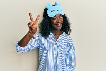 Beautiful african young woman wearing sleep mask and pajama smiling with happy face winking at the...