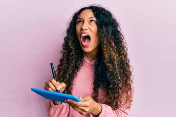 Young latin woman using touchpad writing on screen angry and mad screaming frustrated and furious,...
