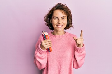 Young brunette woman holding colored pencils smiling with an idea or question pointing finger with...