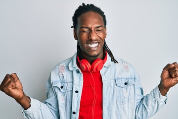 African american man with braids listening to music using headphones very happy and excited doing winner gesture with arms raised, smiling and screaming for success. celebration concept.