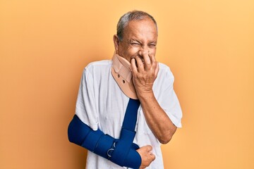 Handsome mature senior man wearing cervical collar and arm on sling laughing and embarrassed giggle covering mouth with hands, gossip and scandal concept