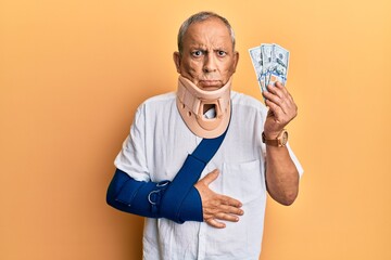 Handsome mature injured man wearing cervical collar and sling holding insurance money skeptic and...
