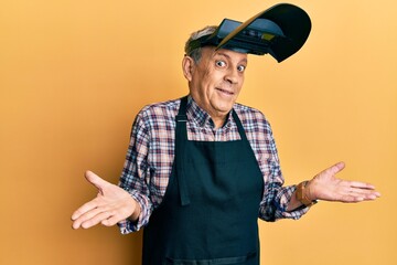 Handsome senior man with grey hair wearing welding protection mask clueless and confused expression...