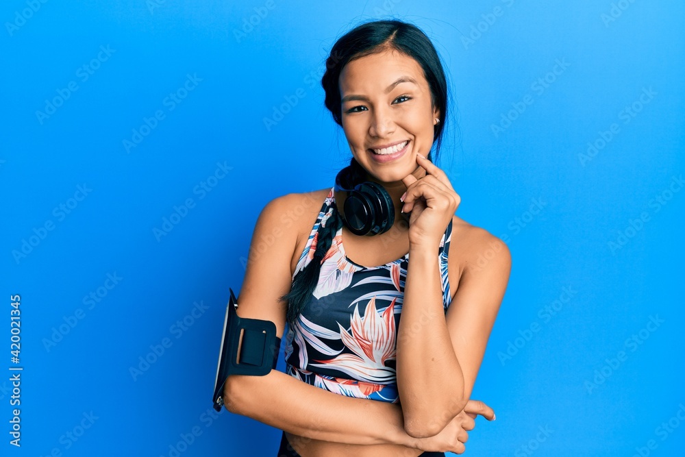 Sticker Beautiful hispanic woman wearing gym clothes and using headphones smiling looking confident at the camera with crossed arms and hand on chin. thinking positive.