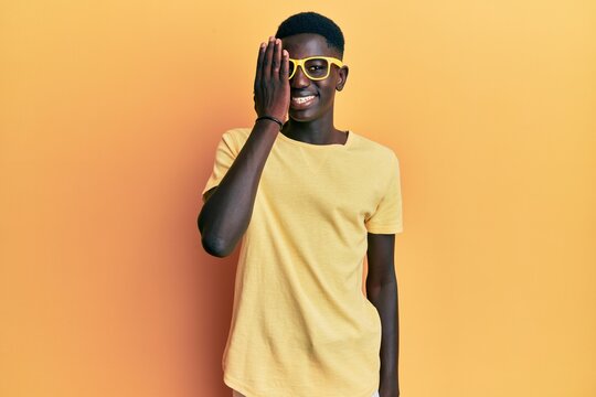 Young african american man wearing casual clothes and glasses covering one eye with hand, confident smile on face and surprise emotion.