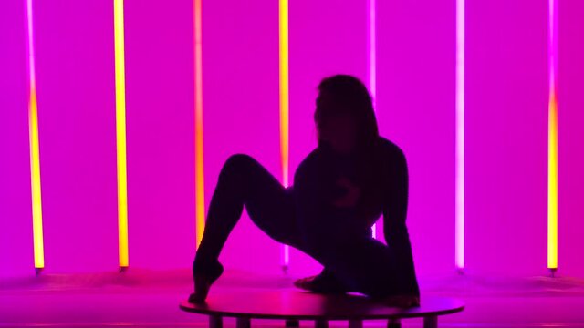 An attractive woman performs a handstand, moves her legs and splits them into a split. Gymnast in the studio against the background of multicolored neon lights. Silhouette. Slow motion. Close up.