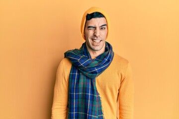 Handsome hispanic man wearing wool hat and winter scarf winking looking at the camera with sexy expression, cheerful and happy face.