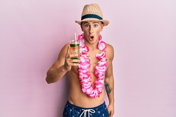 Young caucasian man wearing summer hat and hawaiian lei drinking cocktail scared and amazed with open mouth for surprise, disbelief face - Powered by Adobe