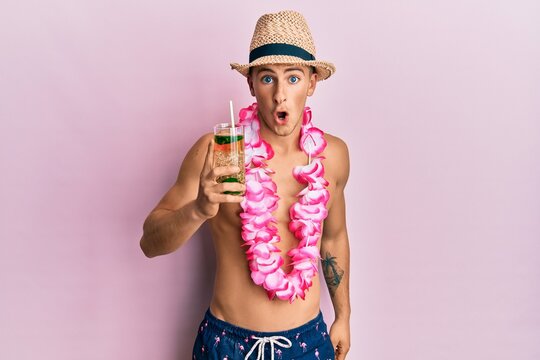 Young caucasian man wearing summer hat and hawaiian lei drinking cocktail scared and amazed with open mouth for surprise, disbelief face