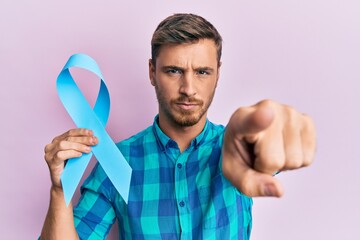 Handsome caucasian man holding blue ribbon pointing with finger to the camera and to you, confident...