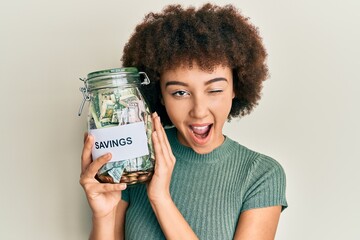 Young hispanic girl holding jar with savings winking looking at the camera with sexy expression,...