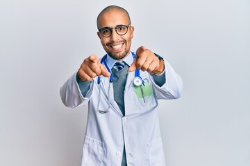 Hispanic adult man wearing doctor uniform and stethoscope pointing to you and the camera with fingers, smiling positive and cheerful