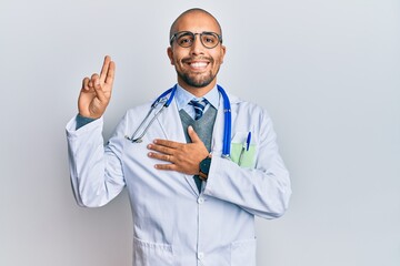 Hispanic adult man wearing doctor uniform and stethoscope smiling swearing with hand on chest and...