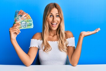 Beautiful blonde young woman holding australian dollars celebrating achievement with happy smile...