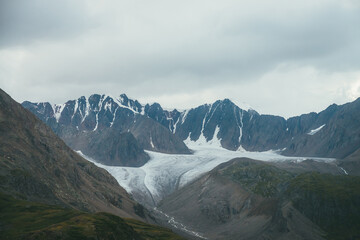 Dramatic alpine landscape with big glacier and mountain river in valley in overcast weather. Atmospheric scenery with great mountain range under cloudy sky. Beautiful mountain ridge under gray sky.