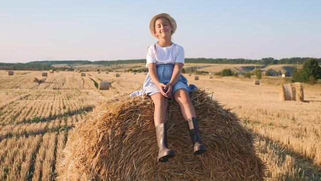 Happy childhood. A happy young girl in a dress and rubber boots is sitting on a haystack in a mown field. Beautiful little girl sitting on a haystack. Fun dream kid concept.