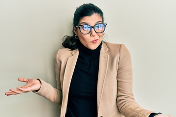 Young hispanic woman wearing business style and glasses clueless and confused expression with arms and hands raised. doubt concept.