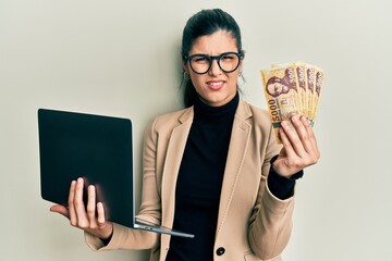 Young hispanic woman wearing business style holding laptop and hungarian forint clueless and...