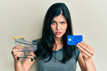 Young hispanic woman holding small supermarket shopping cart and credit card skeptic and nervous, frowning upset because of problem. negative person.