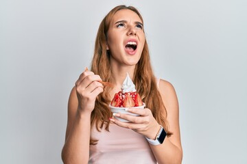 Young blonde woman eating strawberry ice cream angry and mad screaming frustrated and furious, shouting with anger looking up.