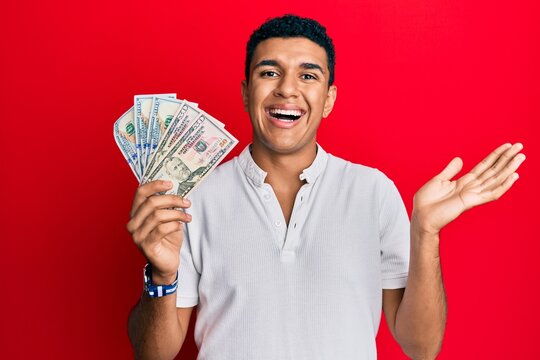 Young arab man holding dollars celebrating achievement with happy smile and winner expression with raised hand