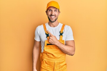 Young handsome man wearing handyman uniform over yellow background cheerful with a smile on face pointing with hand and finger up to the side with happy and natural expression