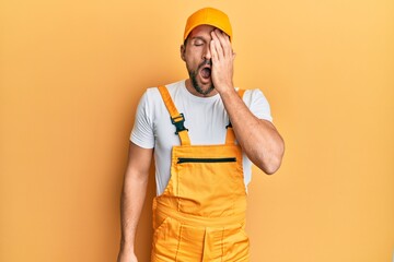 Young handsome man wearing handyman uniform over yellow background yawning tired covering half face, eye and mouth with hand. face hurts in pain.