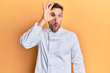 Fototapeta premium Handsome man with beard wearing professional cook uniform doing ok gesture shocked with surprised face, eye looking through fingers. unbelieving expression.