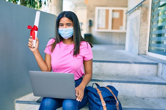 Young latin student girl wearing medical mask using laptop and holding diploma at university.