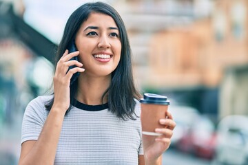 Young hispanic woman talking on the smartphone and drinking take away coffee at the city.