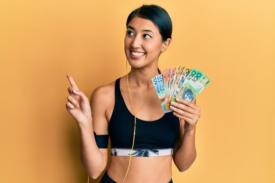 Beautiful asian young sport woman holding australian dollars smiling happy pointing with hand and finger to the side