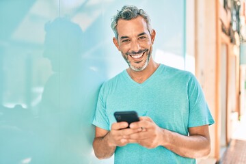 Middle age grey-haired man using smartphone leaning on the wall at city.