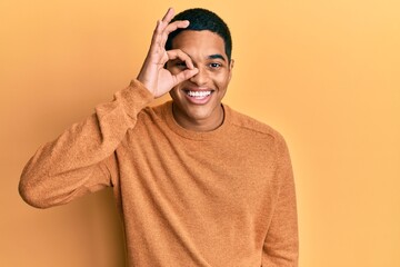 Young handsome hispanic man wearing casual winter sweater doing ok gesture with hand smiling, eye looking through fingers with happy face.