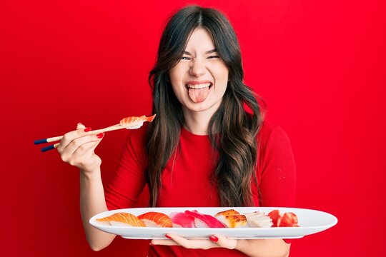 Young beautiful caucasian girl eating sushi using chopsticks sticking tongue out happy with funny expression.