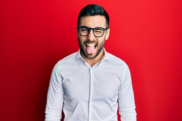Young hispanic businessman wearing shirt and glasses sticking tongue out happy with funny expression. emotion concept.