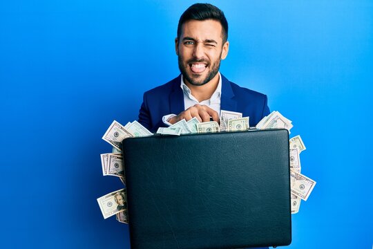Young hispanic businessman holding briefcase full of dollars sticking tongue out happy with funny expression.
