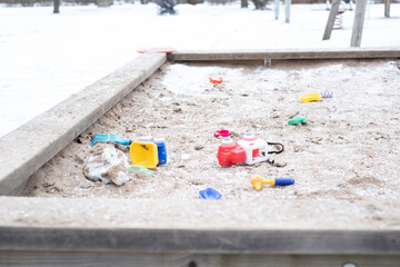 Abandoned colorful toys in sandbox at empty public playground. Kids toys scattered around in snowy frozen sandbox during winter lockdown.