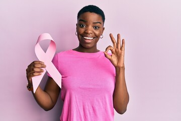 Young african american woman holding pink cancer ribbon doing ok sign with fingers, smiling...