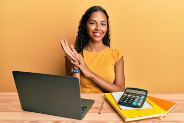 Young african american girl working at the office with laptop and calculator clapping and...