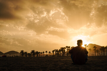 Hombre de espaldas sentado en la playa mirando puesta de Sol en Carboneras, Almería, Andalucía,...