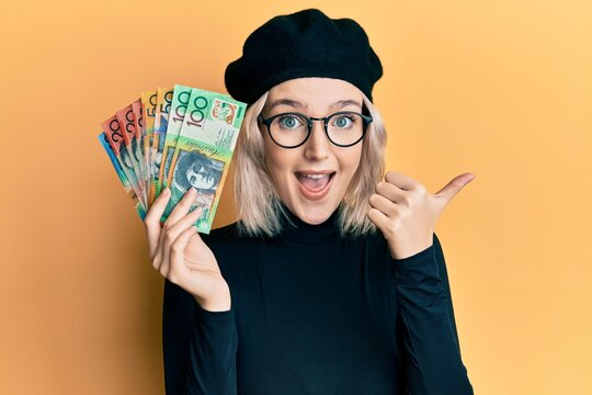 Young blonde girl holding australian dollars pointing thumb up to the side smiling happy with open mouth
