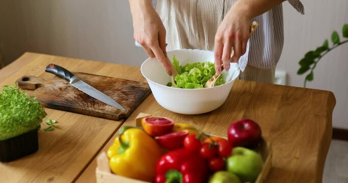 Woman is preparing vegetable salad in the kitchen, mixing leaf of salad and vegetables ina white bowl, Healthy food concept, vegan or diet.